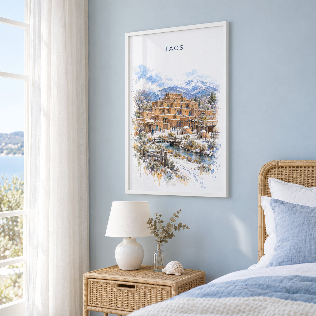 Taos watercolor print in a white frame on a pale blue wall beside a rattan bed in a coastal bedroom