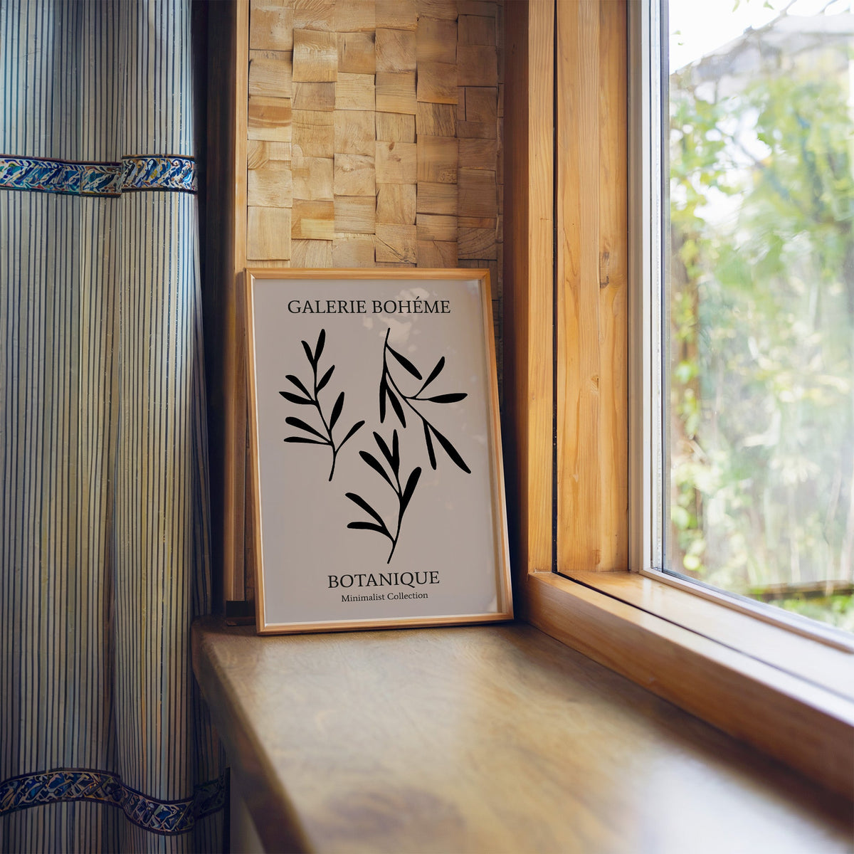 Three black sprigs on taupe abstract botanical print in a light wood frame leaning on a wooden window sill by a garden view