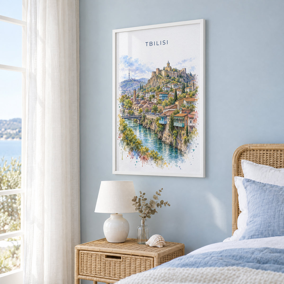 Tbilisi watercolor print in a white frame on a pale blue wall beside a rattan bed in a coastal bedroom