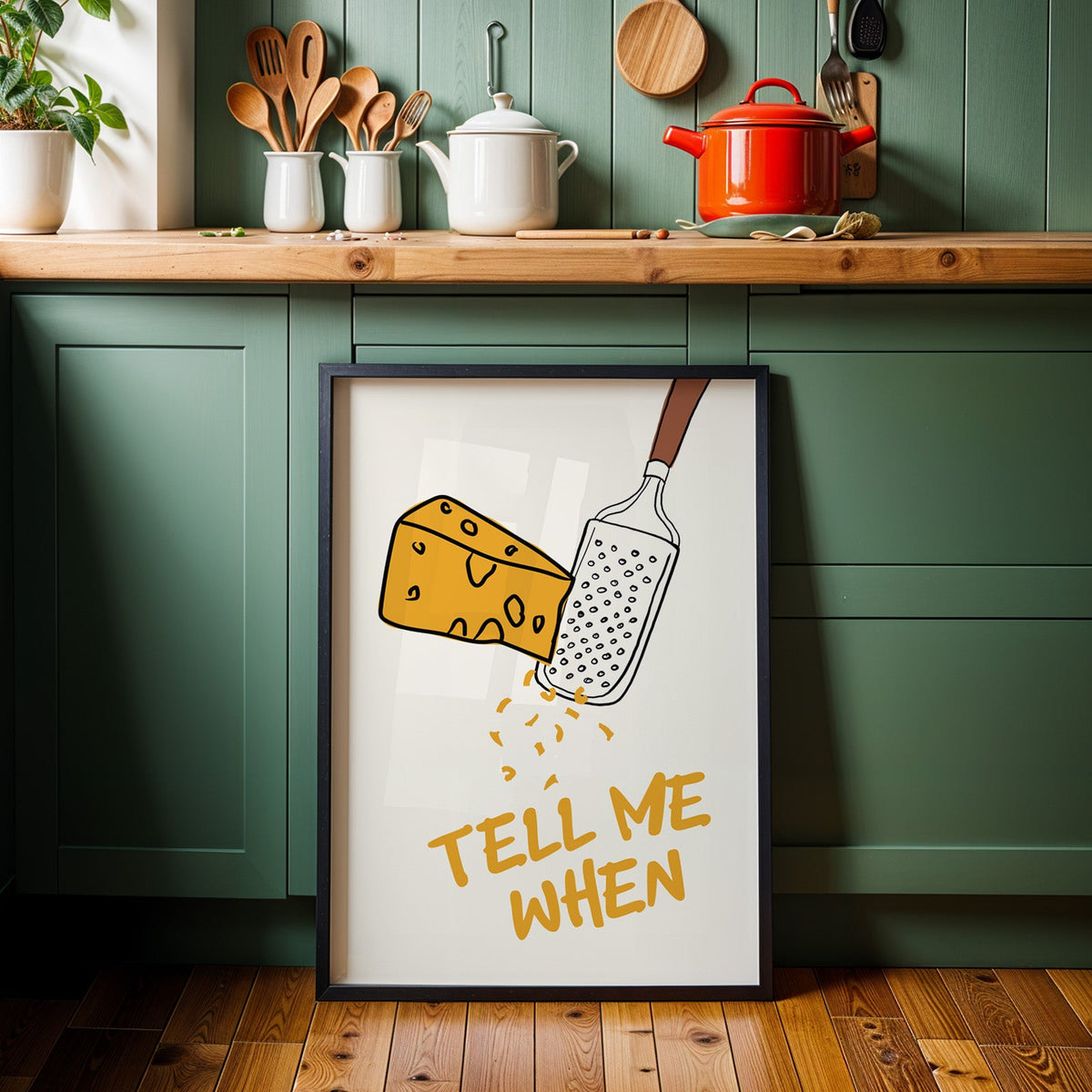 Tell me when cheese grater print in a black frame against sage green kitchen cabinets