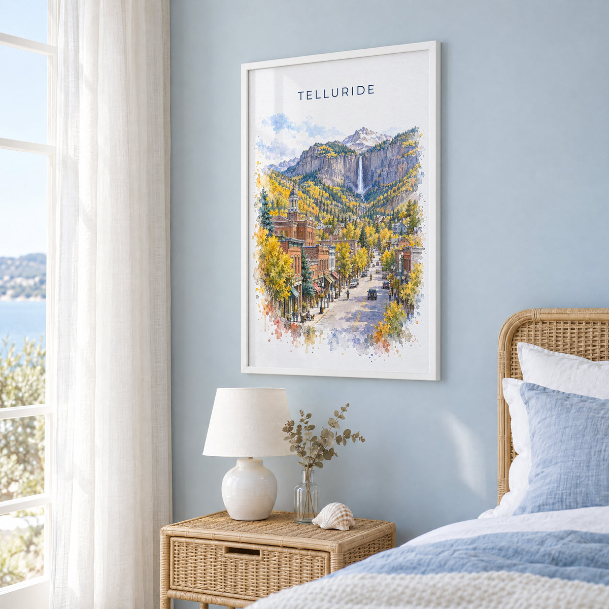 Telluride watercolor print in a white frame on a pale blue wall beside a rattan bed in a coastal bedroom