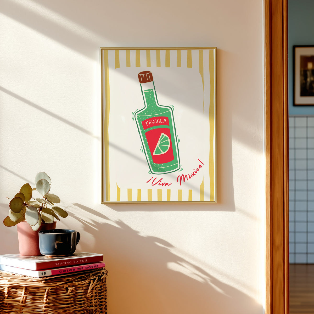 Tequila bottle print in a gold frame above a basket of books and a pilea plant