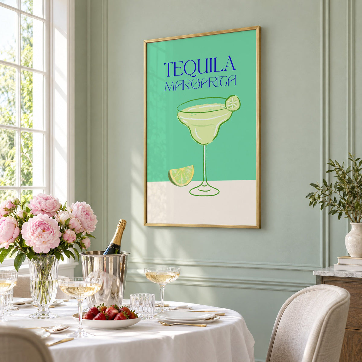TEQUILA MARGARITA green glass and lime print in a brass frame on a sage wall above a brunch table with champagne and peonies, seen at an angle