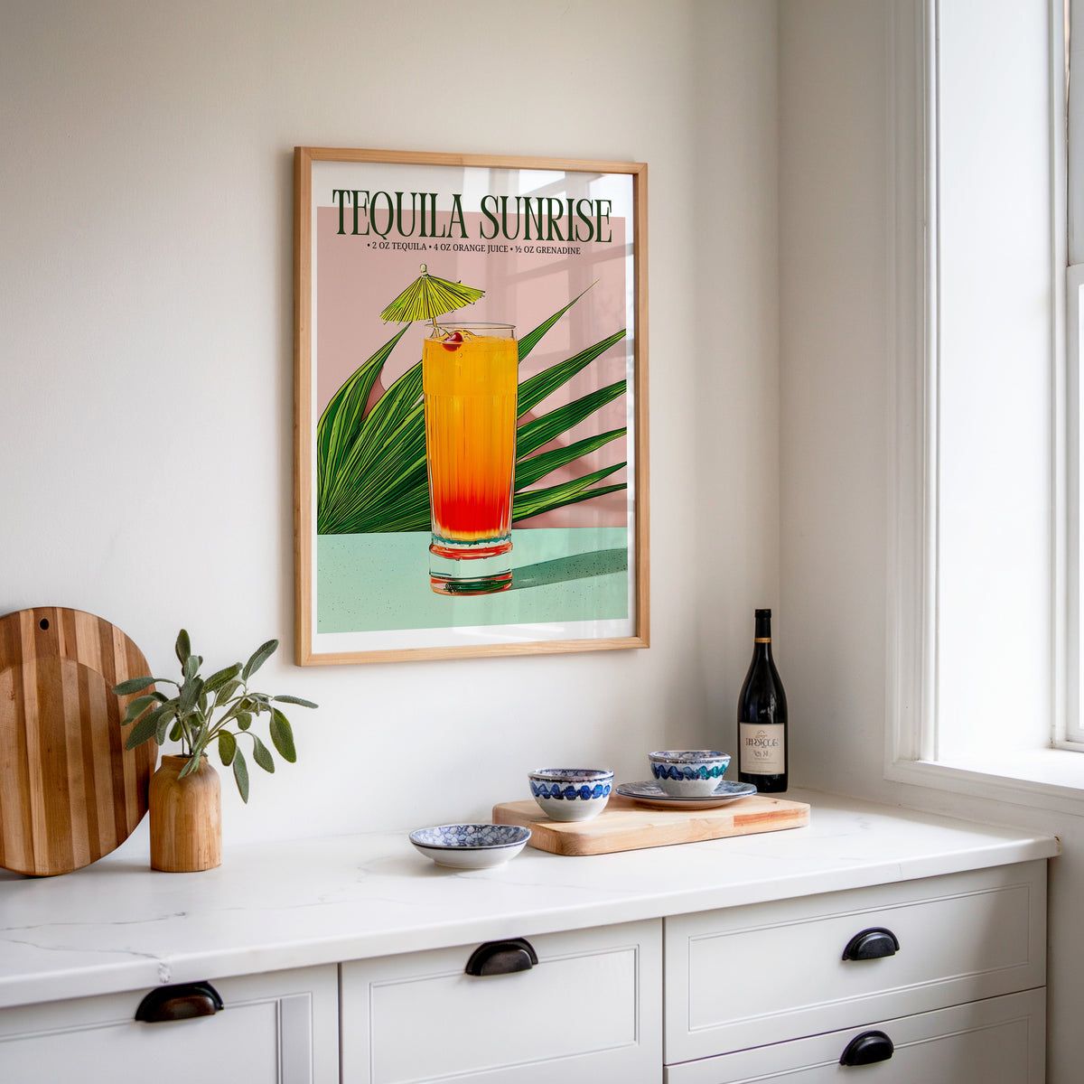 Tequila sunrise recipe print in an oak frame above a white sideboard with a bottle of wine
