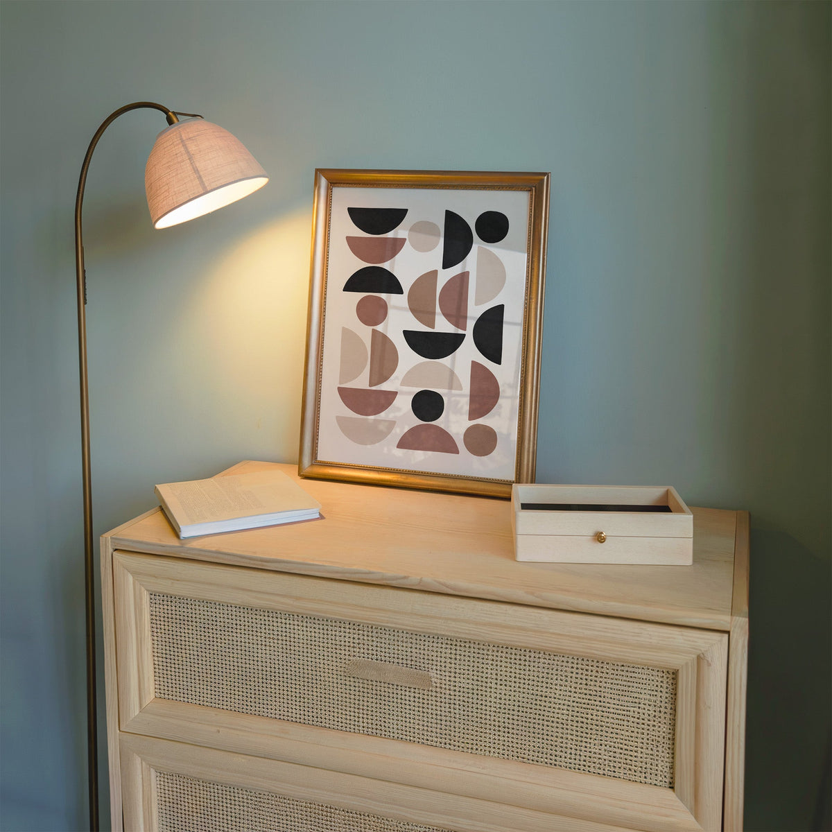 Terracotta and black half circle mosaic print shown in a gold frame on a cane dresser lit by a floor lamp