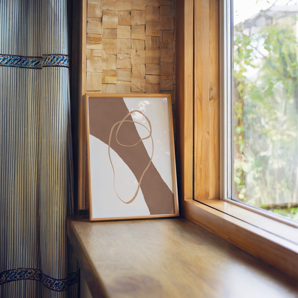 Terracotta diagonal band and loop line print in a light wood frame leaning on a wooden window sill by a garden view