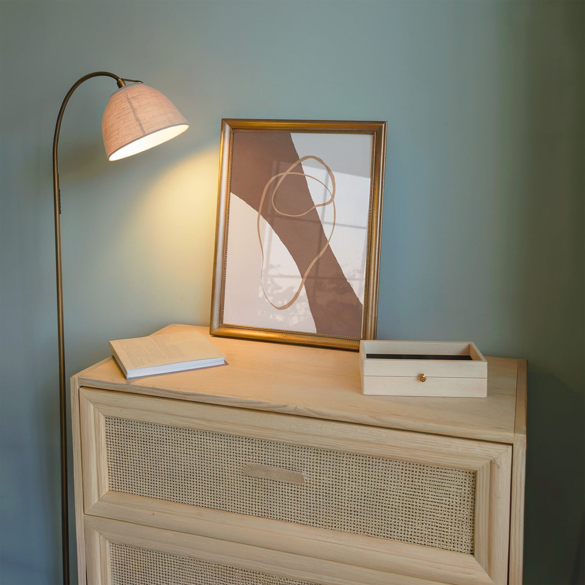 Terracotta diagonal band and loop line print shown in a gold frame on a cane dresser lit by a floor lamp