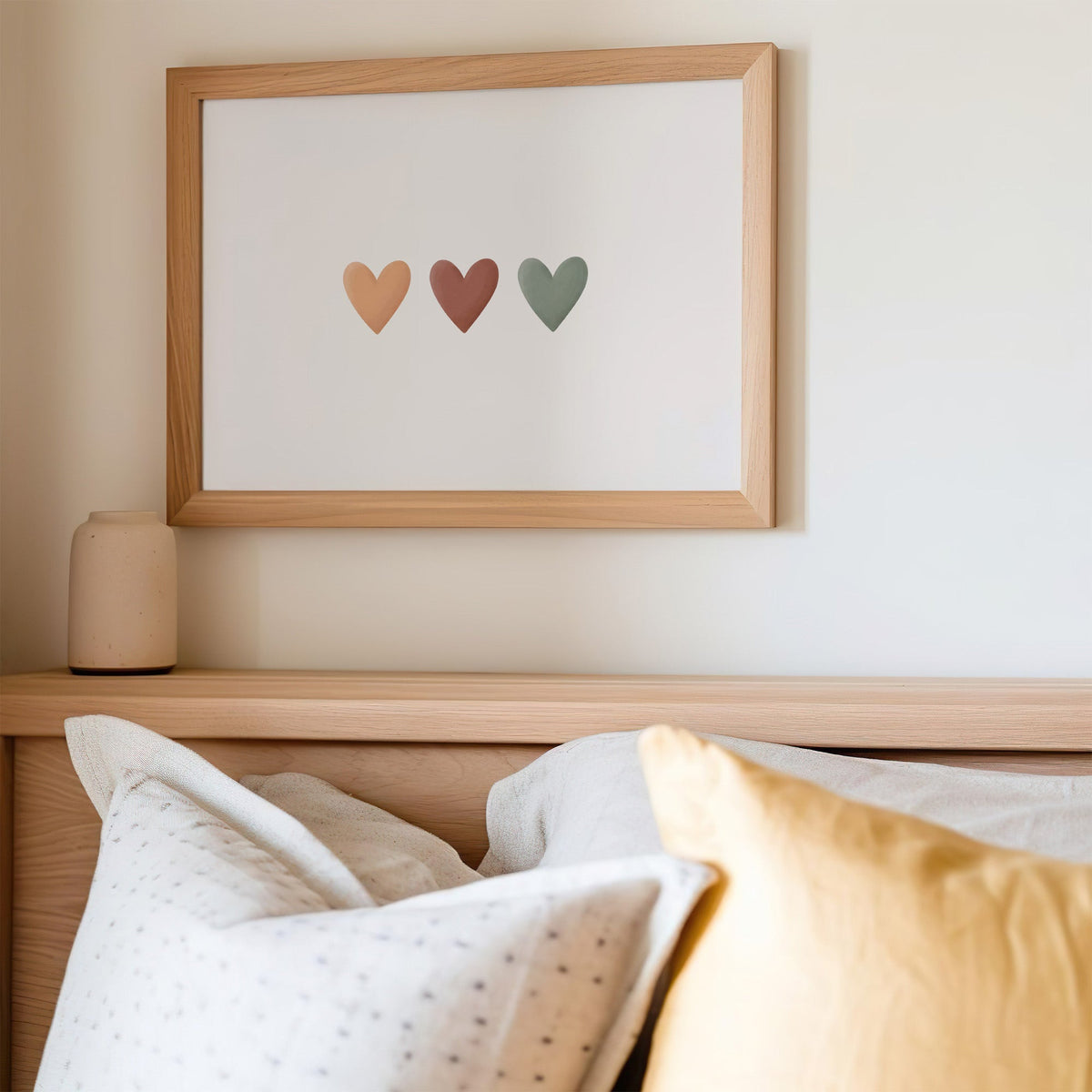 Three peach, terracotta and sage hearts in a row nursery print in an oak frame above a wooden headboard with a mustard pillow