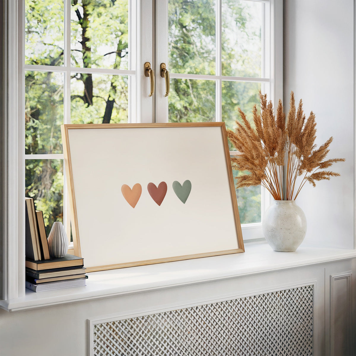 Three peach, terracotta and sage hearts in a row nursery print in an oak frame on a white windowsill beside dried pampas grass