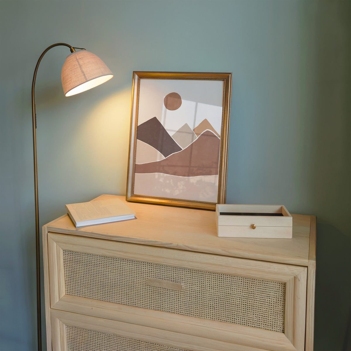 Terracotta sun over earth-tone mountains boho print shown in a gold frame on a cane dresser lit by a floor lamp