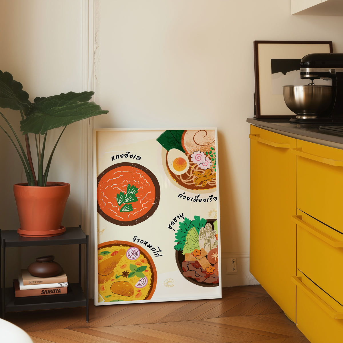 Thai food bowls print in a white frame leaning on parquet beside a bright yellow kitchen