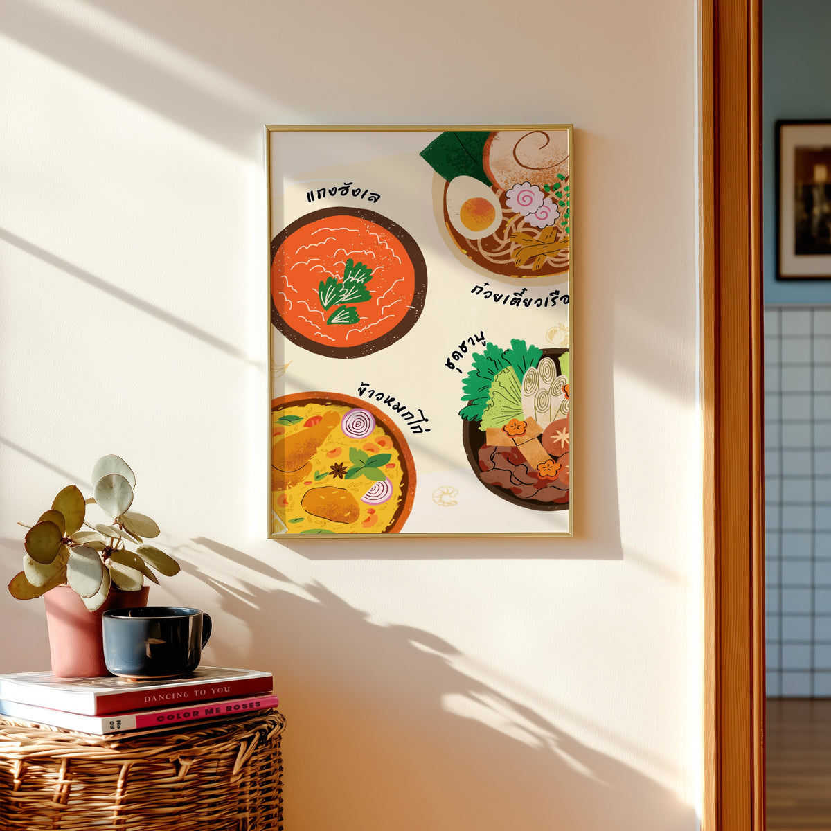 Thai food bowls print shown in a gold frame above a basket of books and a pilea plant