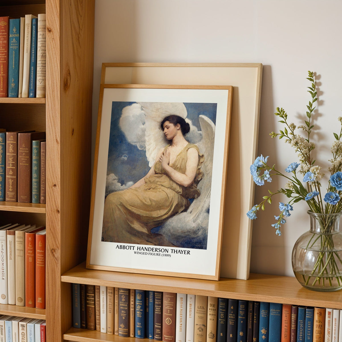 Winged figure print in an oak frame leaning on a bookshelf beside blue flowers