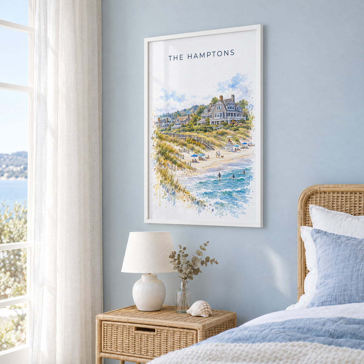 The Hamptons watercolor print in a white frame on a pale blue wall beside a rattan bed in a coastal bedroom