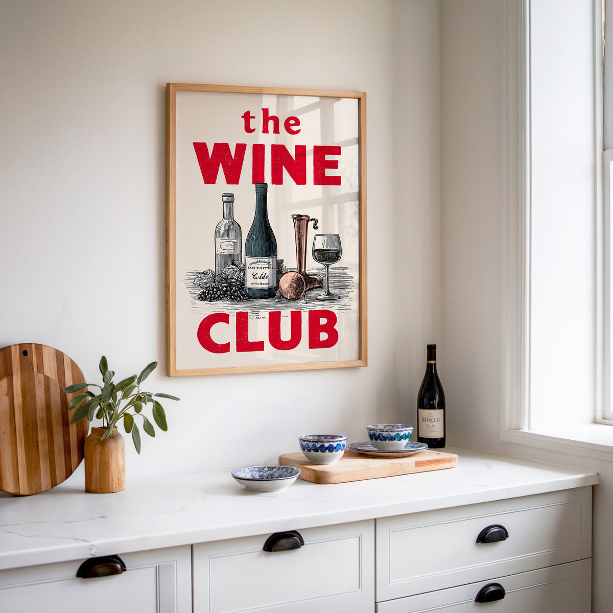 The Wine Club print in an oak frame above a white sideboard with a bottle of wine