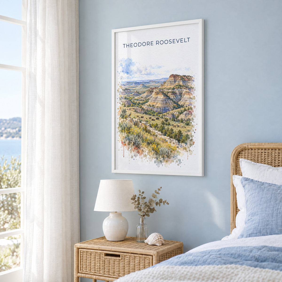 Theodore Roosevelt National Park watercolor in a white frame on a pale blue wall beside a rattan bed in a coastal bedroom