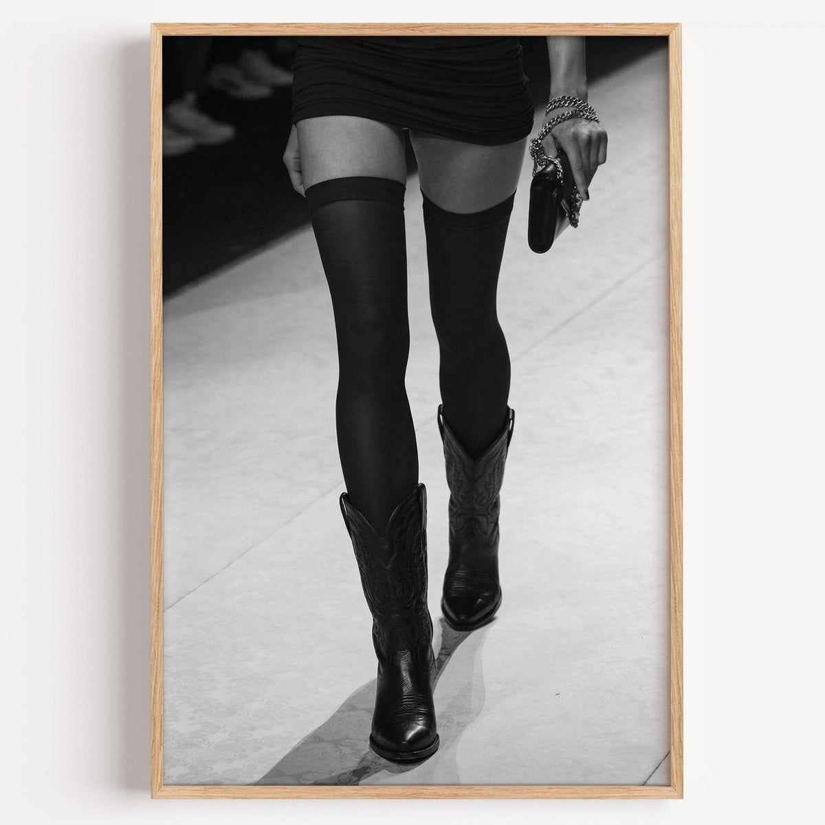 Legs in stockings and cowboy boots on runway print in a light oak frame on a plain white wall