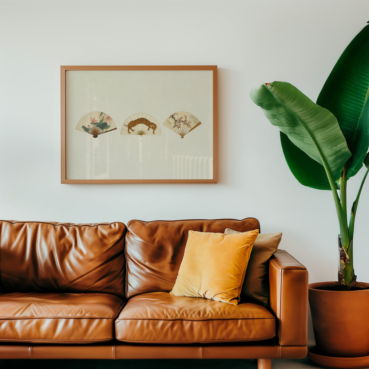 Three painted folding fans print in a warm oak frame above a cognac leather sofa with a mustard pillow