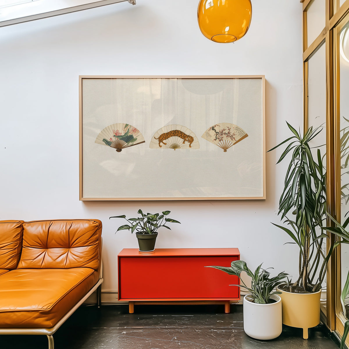 Three painted folding fans print in a light wood frame above a red cabinet beside a tan leather daybed