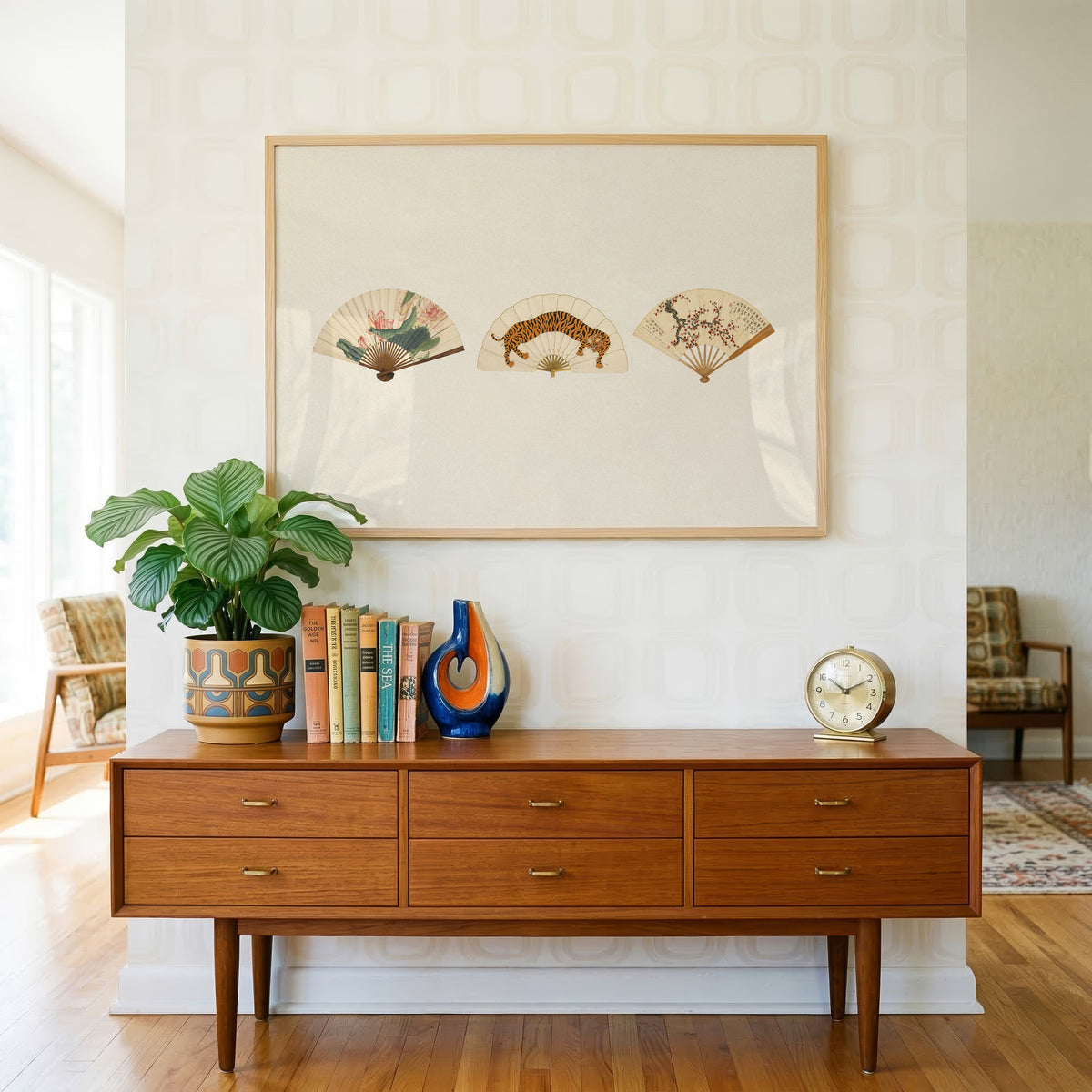 Three painted folding fans print in a light oak frame above a teak credenza with books and a brass clock