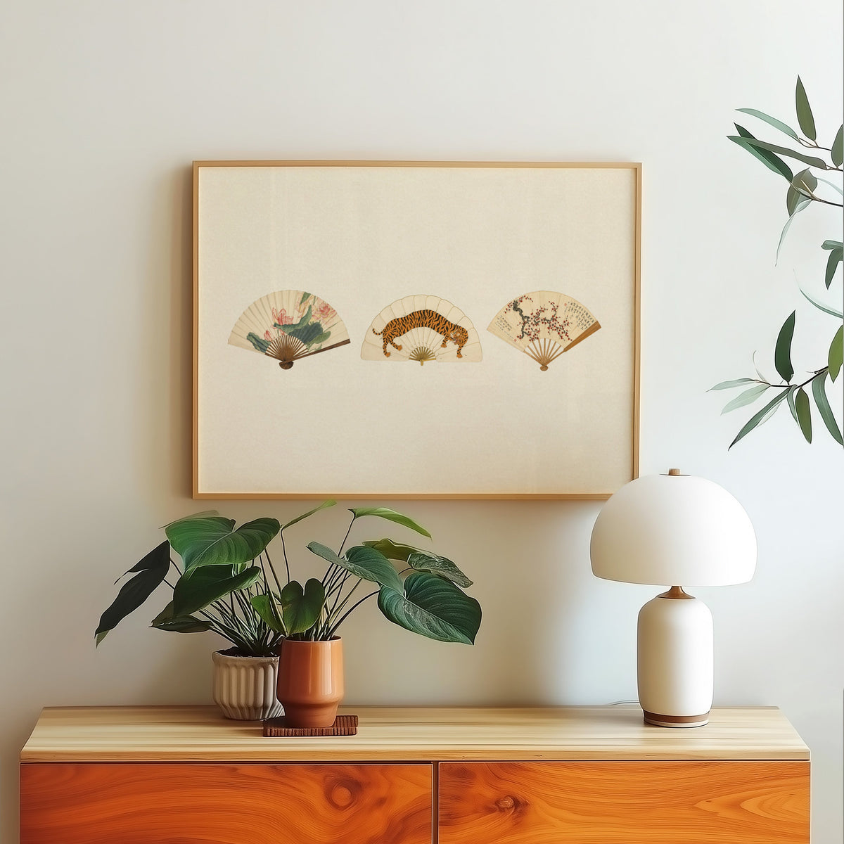 Three painted folding fans print in a light oak frame above a wood sideboard with a white dome lamp