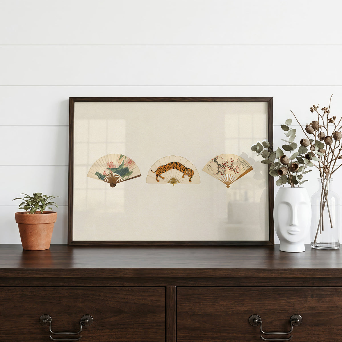 Three painted folding fans print in a dark walnut frame on a dark wood dresser beside a white face vase