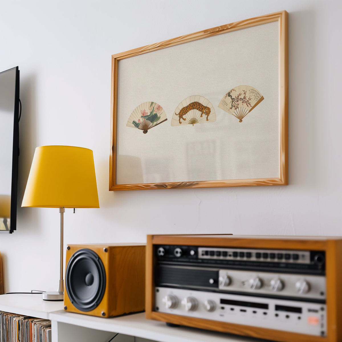 Three painted folding fans print in a pine frame above a vintage stereo and a yellow table lamp