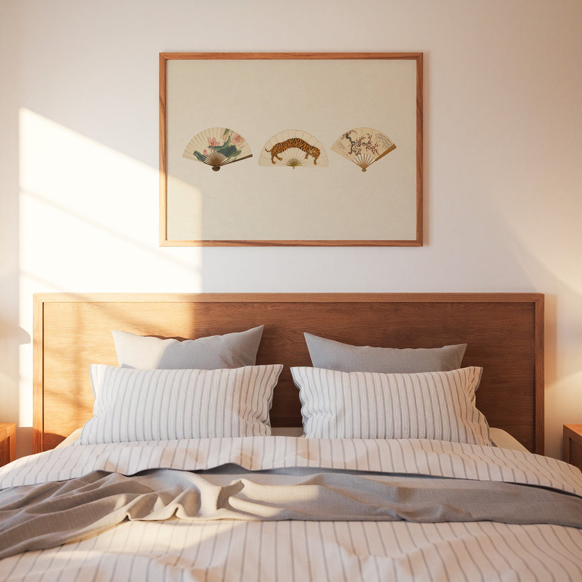 Three painted folding fans print in a warm wood frame above a bed with striped linen bedding