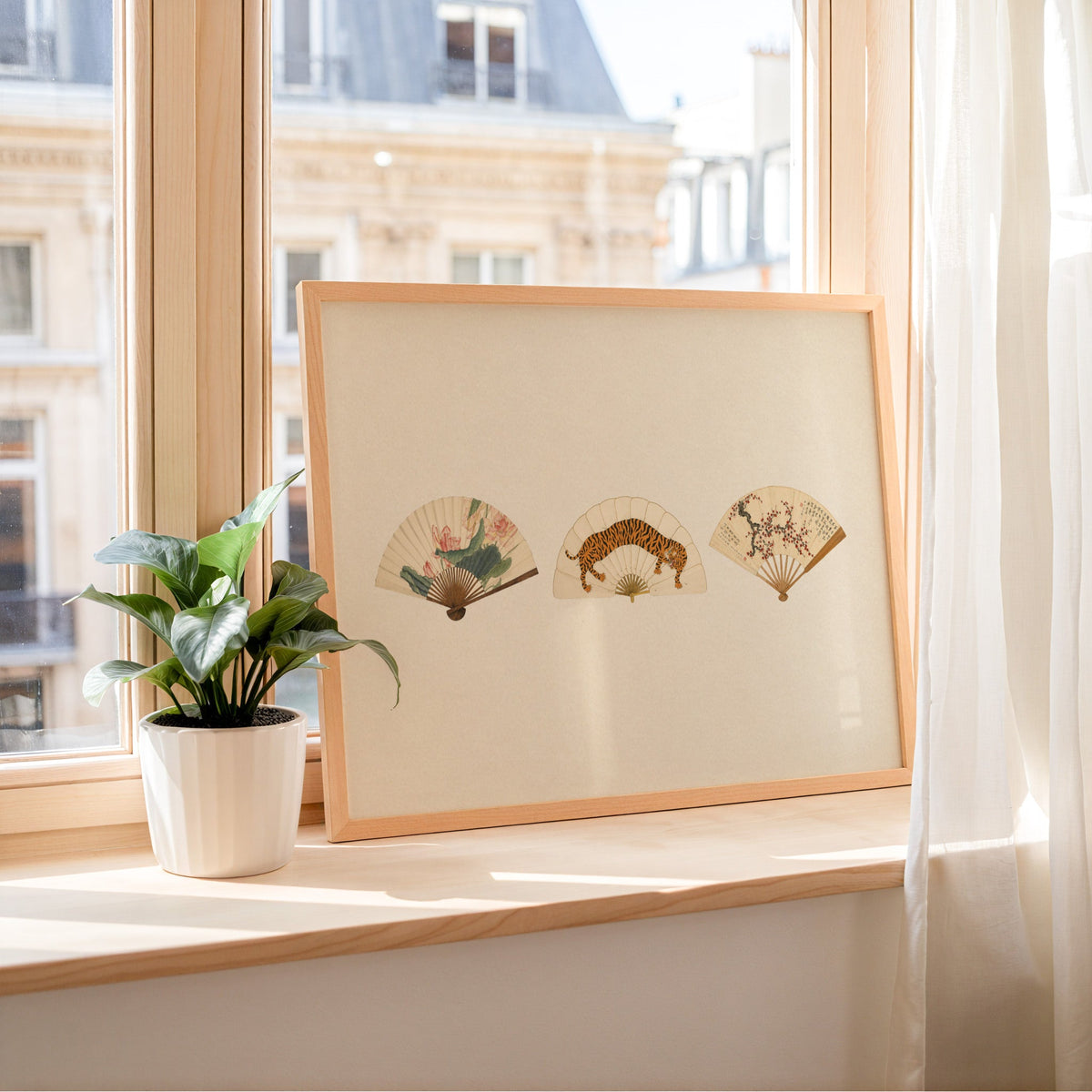 Three painted folding fans print in a light wood frame leaning on a sunny windowsill by sheer curtains