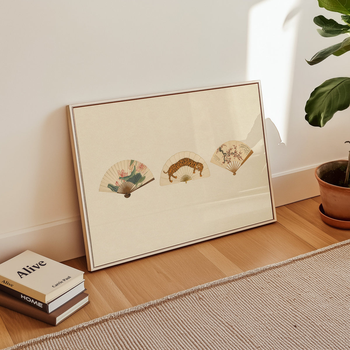 Three painted folding fans print in a white floater frame leaning on a wood floor beside stacked books