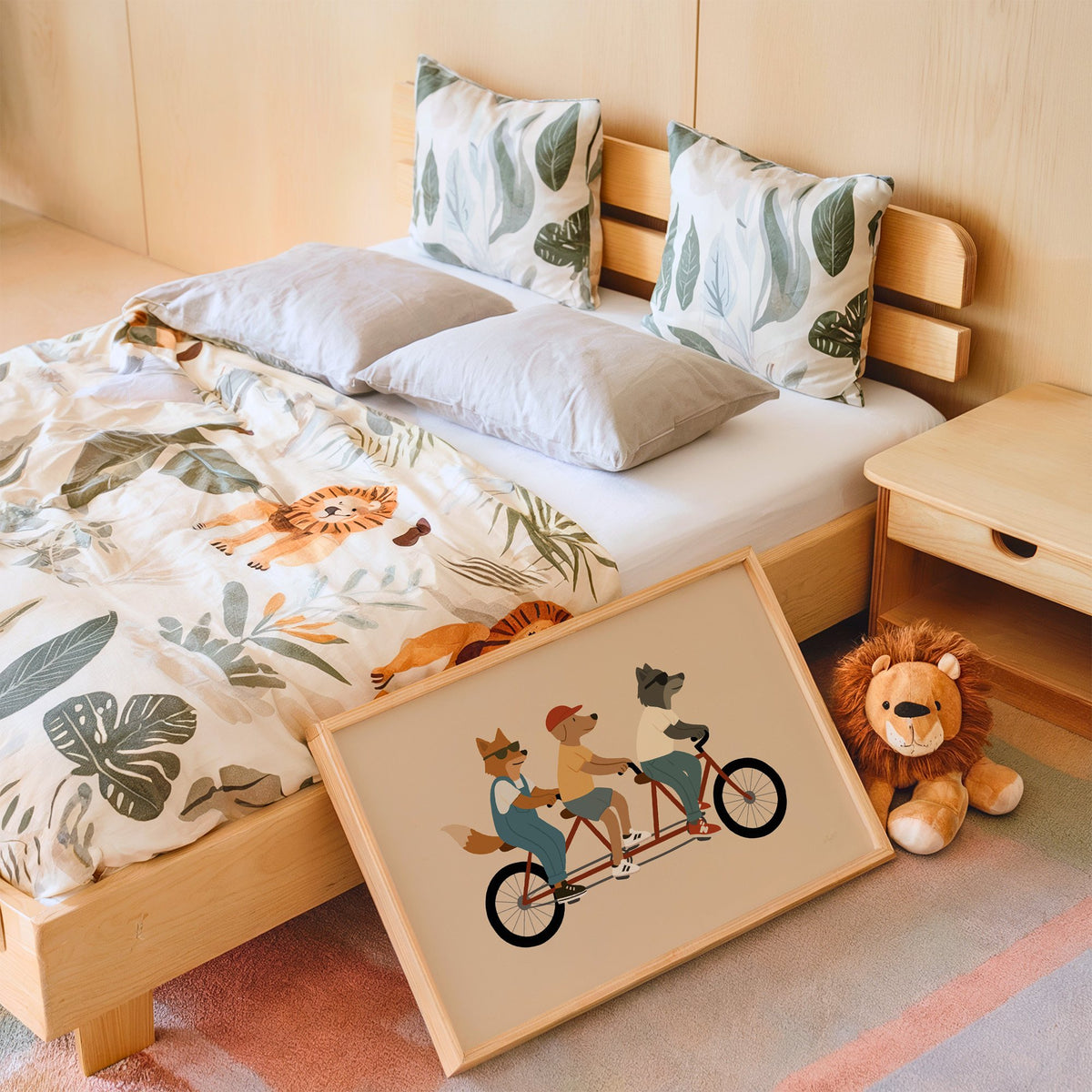 Animal friends riding tandem bike nursery print in an oak frame leaning on a kids bed by a lion plush