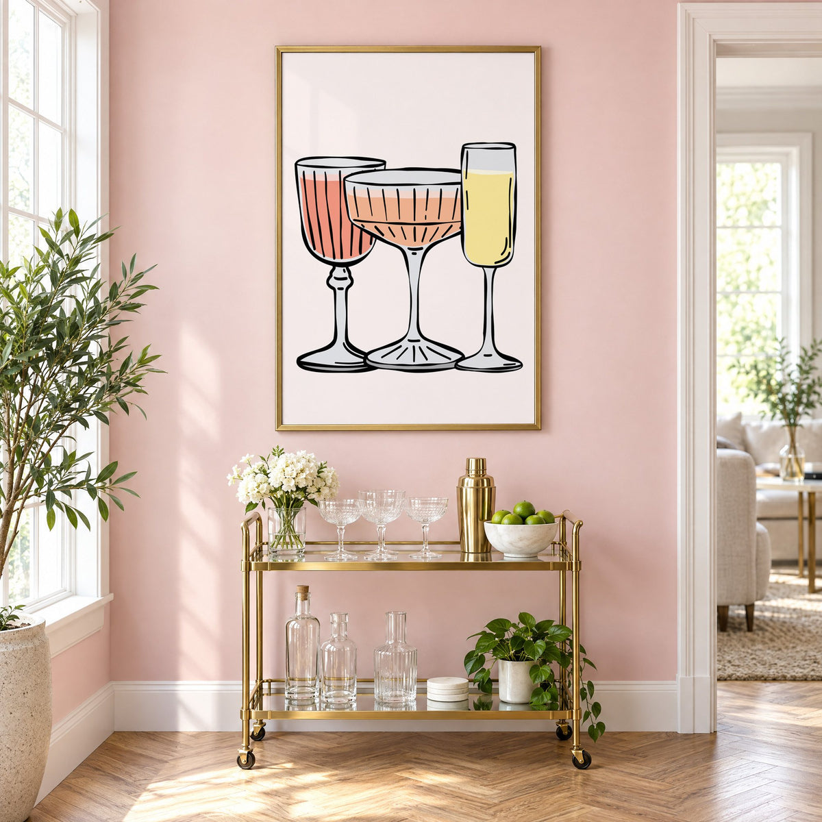 Three pastel cocktail glasses and champagne flute print in a brass frame on a blush pink wall above a gold bar cart with glassware
