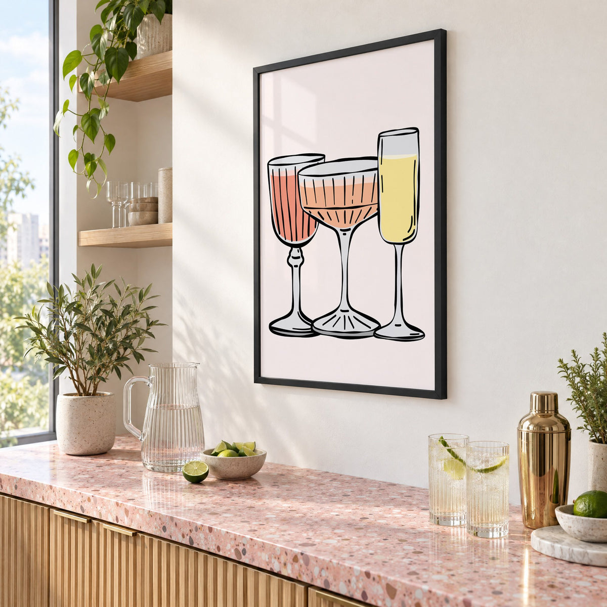 Three pastel cocktail glasses and champagne flute print in a black frame above a pink terrazzo cocktail counter with limes, seen at an angle