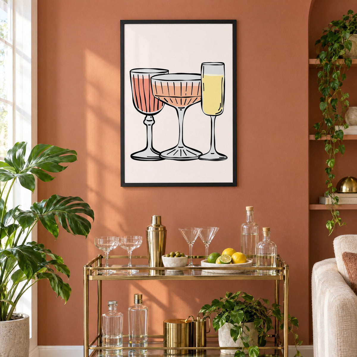 Three pastel cocktail glasses and champagne flute print in a black frame on a terracotta wall above a brass bar cart with coupes and citrus