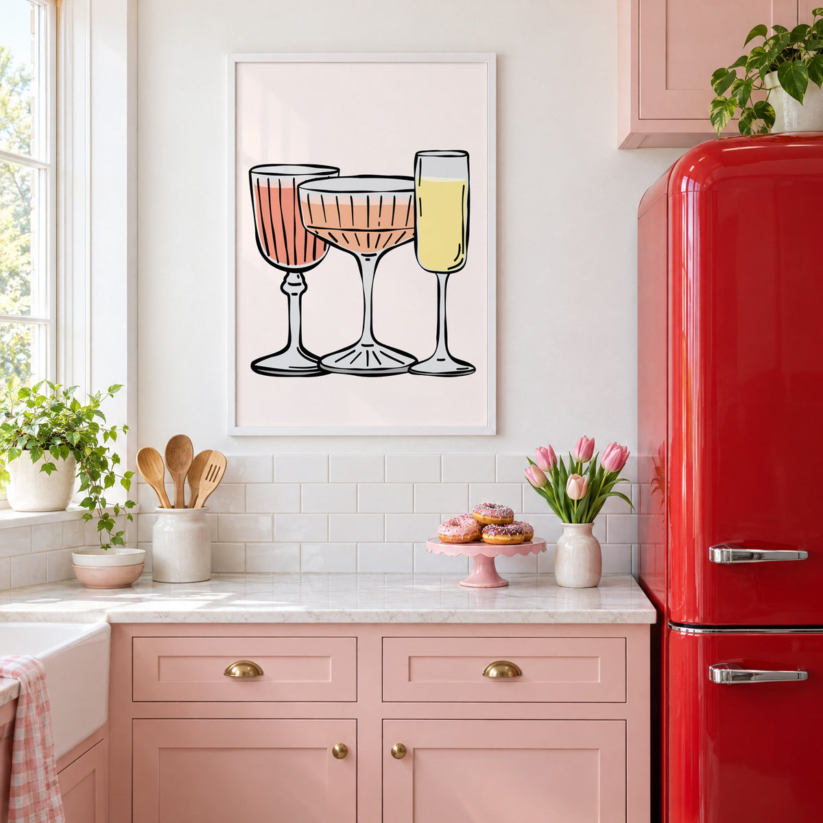 Three pastel cocktail glasses and champagne flute print in a white frame above pink cabinets beside a red retro fridge