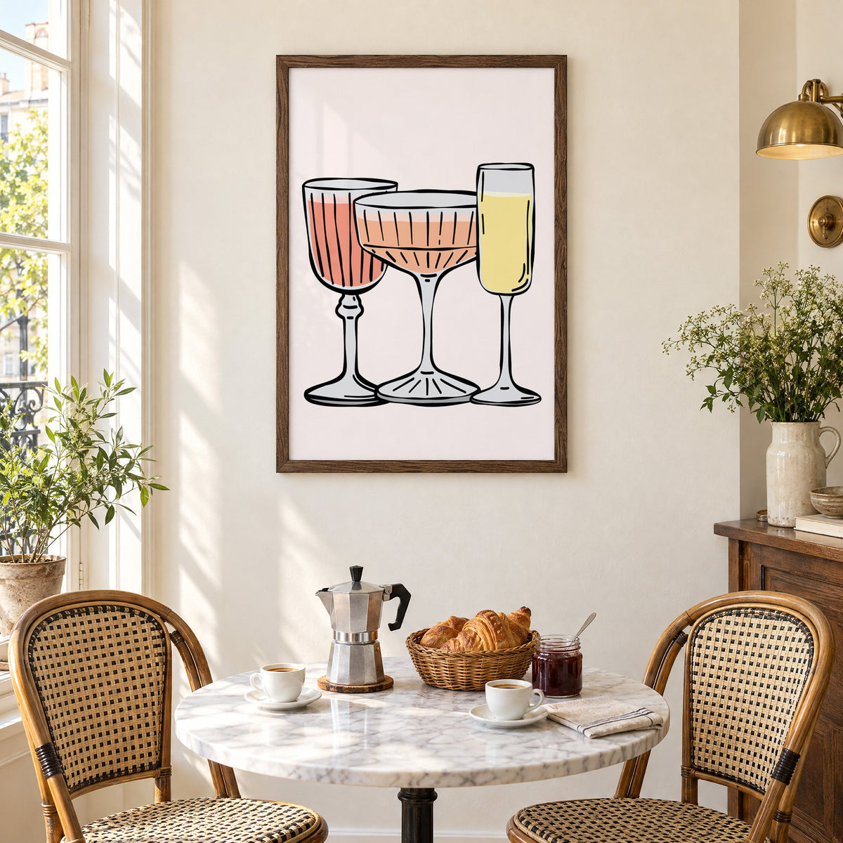 Three pastel cocktail glasses and champagne flute print in a walnut frame above a marble bistro table with croissants and rattan chairs