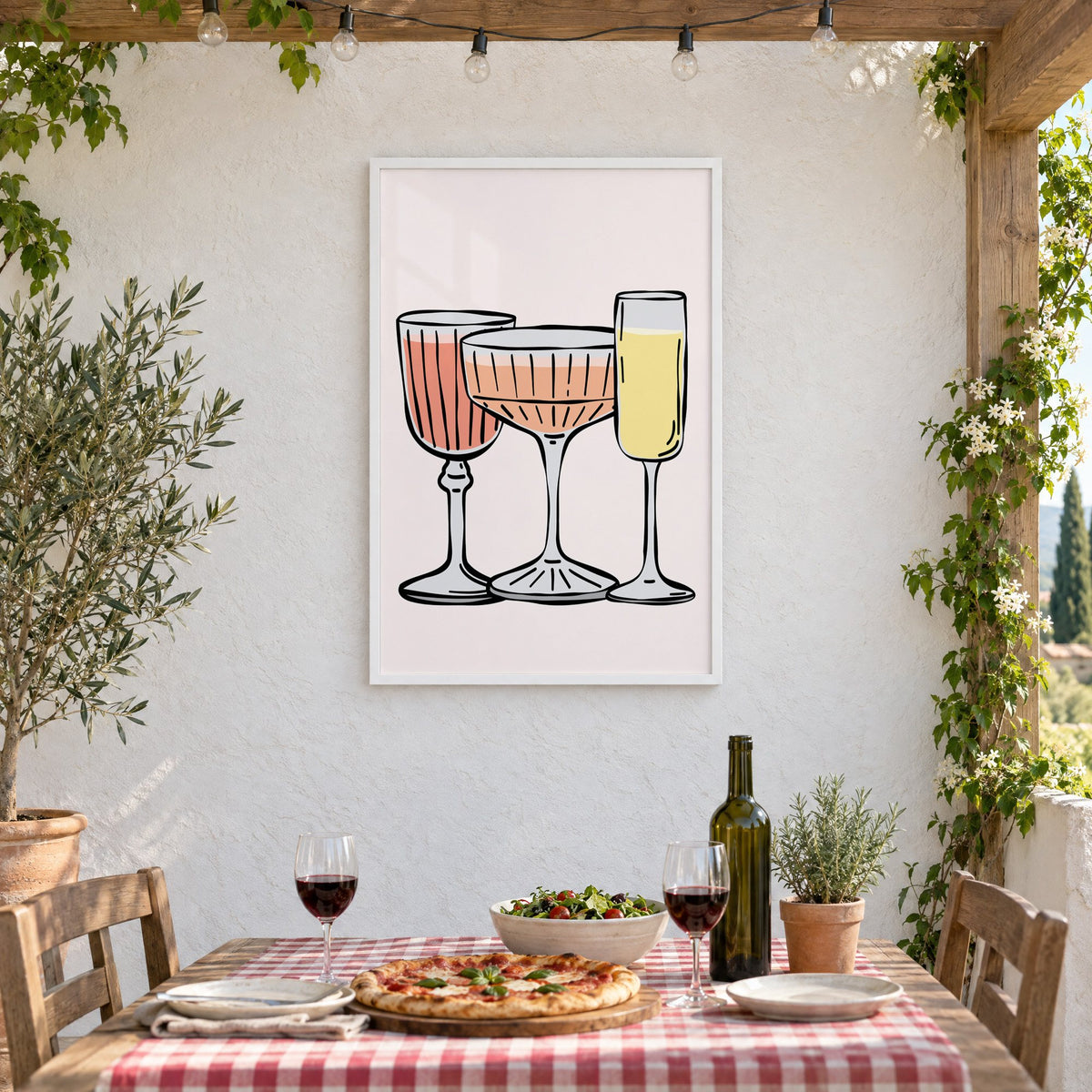 Three pastel cocktail glasses and champagne flute print in a white frame on a stucco wall above a checked-cloth table with pizza and red wine