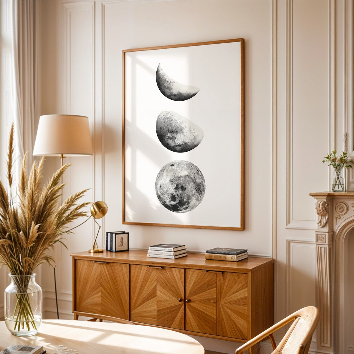 Three moon phases stacked vertically on white print in an oak frame above a sunburst wood sideboard with pampas grass