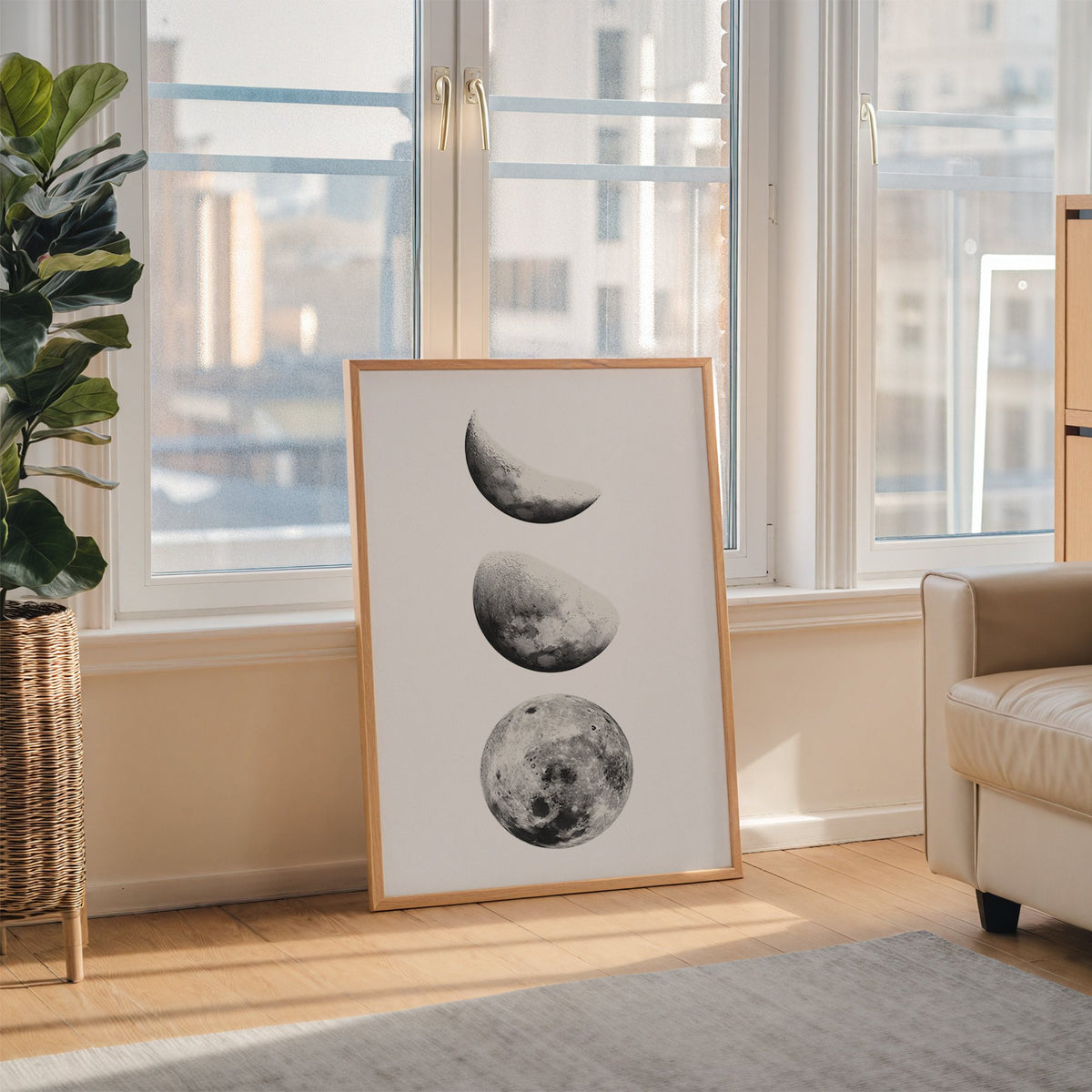 Three moon phases stacked vertically on white print in an oak frame leaning on the floor beside a fiddle leaf fig