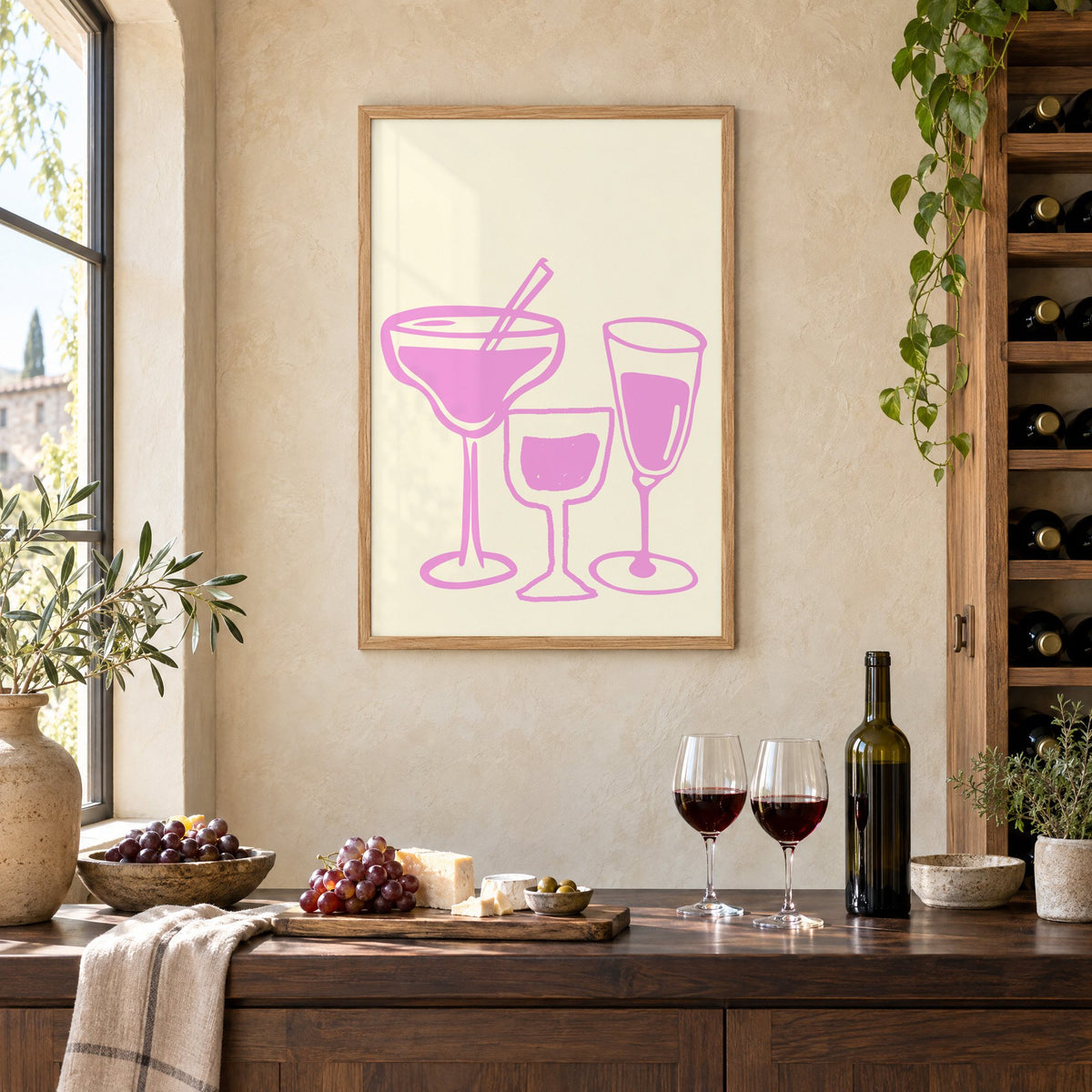 Three pink cocktail glasses linocut-style print in an oak frame on a limewashed wall above a wine counter with red wine, cheese and grapes