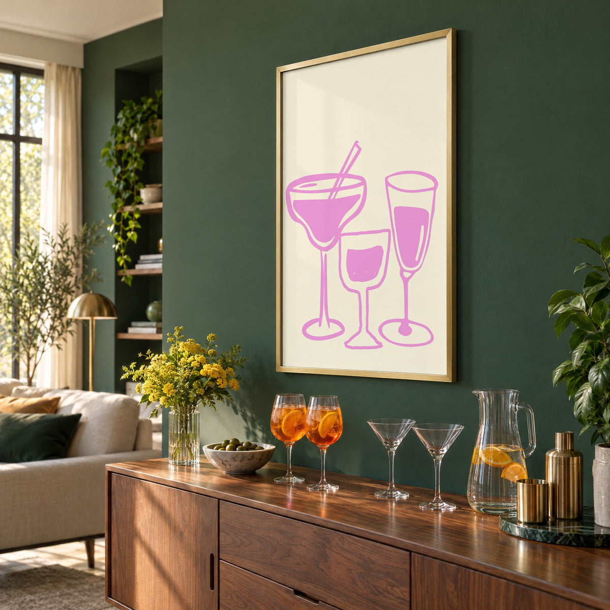 Three pink cocktail glasses linocut-style print in a brass frame on a bottle green wall above a sideboard with spritz and martini glasses, seen at an angle