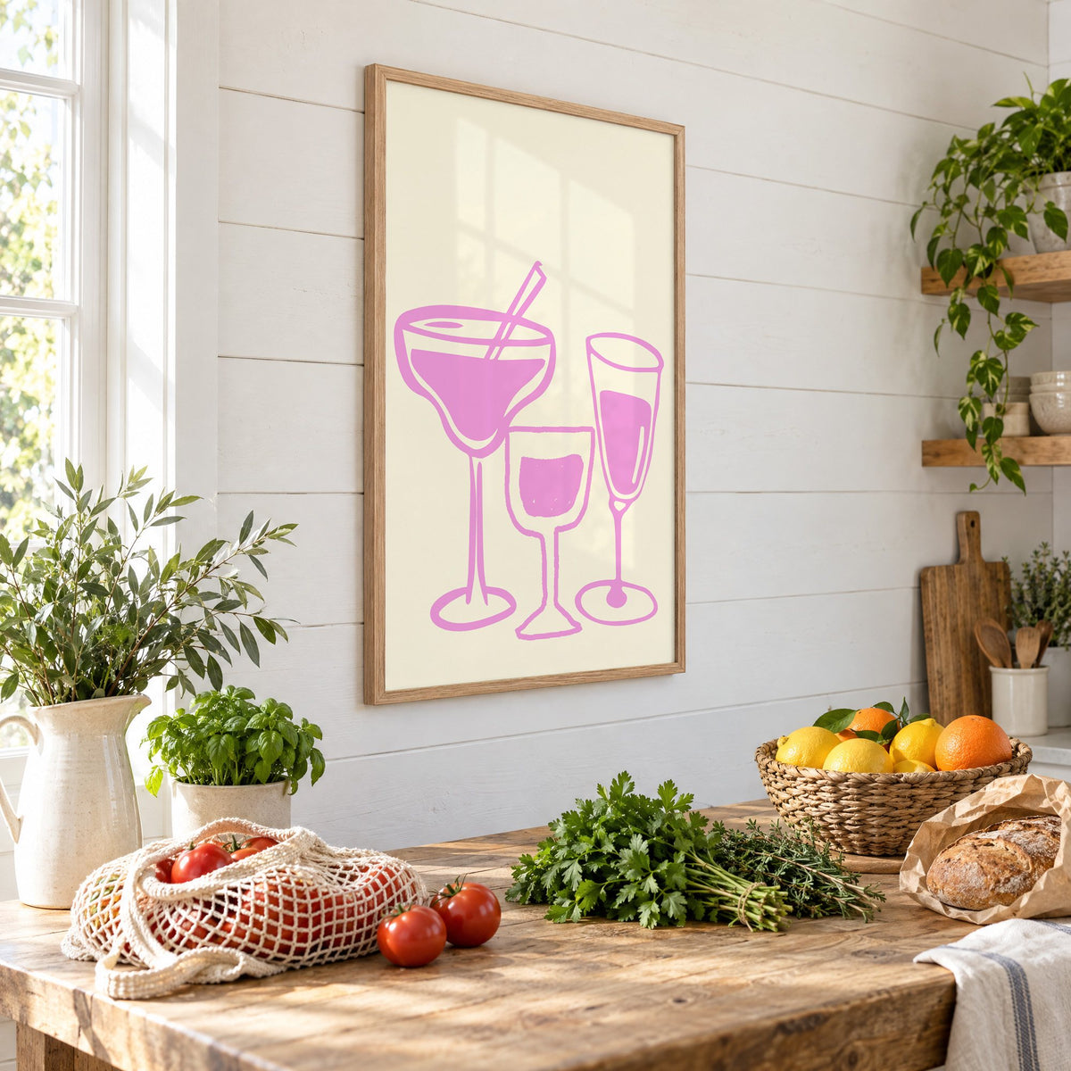 Three pink cocktail glasses linocut-style print in an oak frame on a shiplap wall above a farm table with market tomatoes and citrus, seen at an angle