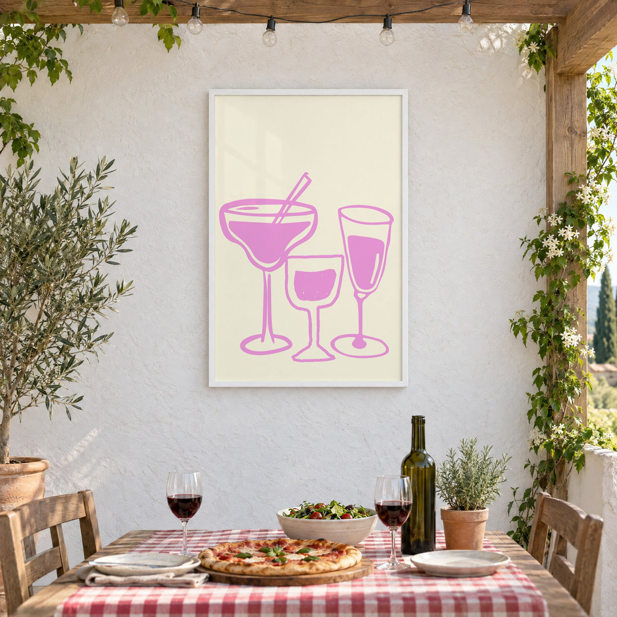 Three pink cocktail glasses linocut-style print in a white frame on a stucco wall above a checked-cloth table with pizza and red wine