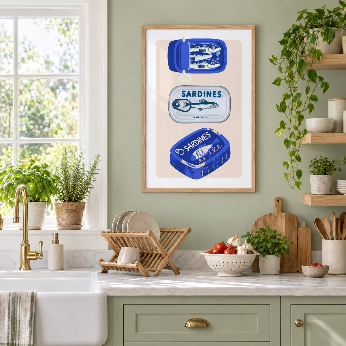 Three sardine tins cobalt print in an oak frame on a sage green kitchen wall beside a farmhouse sink and herbs