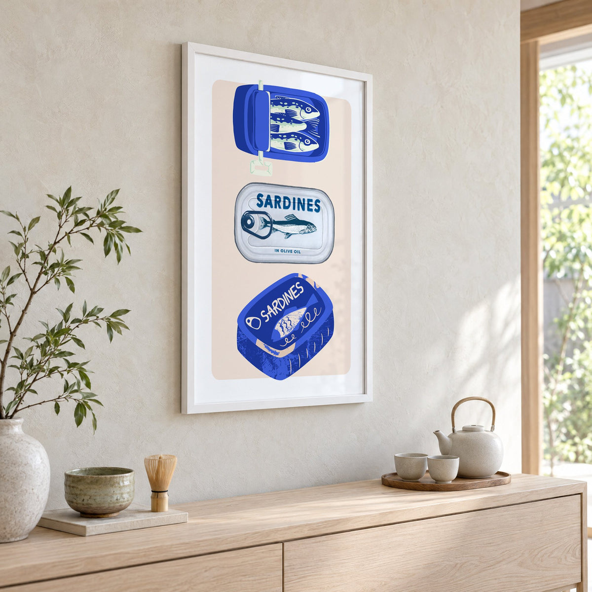 Three sardine tins cobalt print in a white frame above a pale wood console with a matcha tea set