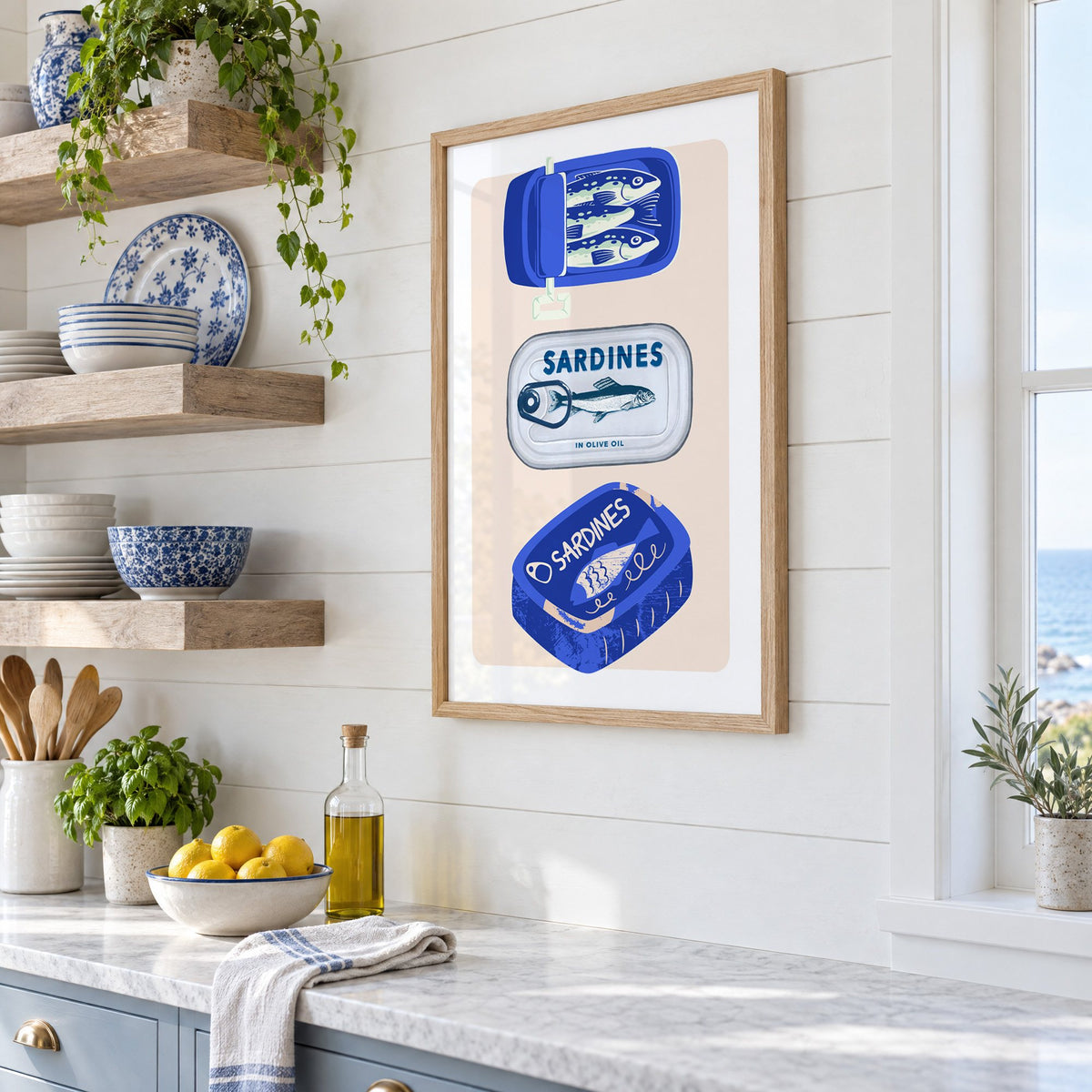 Three sardine tins cobalt print in an oak frame in a white coastal kitchen beside blue and white plates
