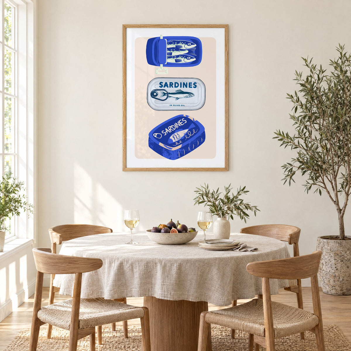 Three sardine tins cobalt print in an oak frame above a round linen-covered dining table with figs and wine