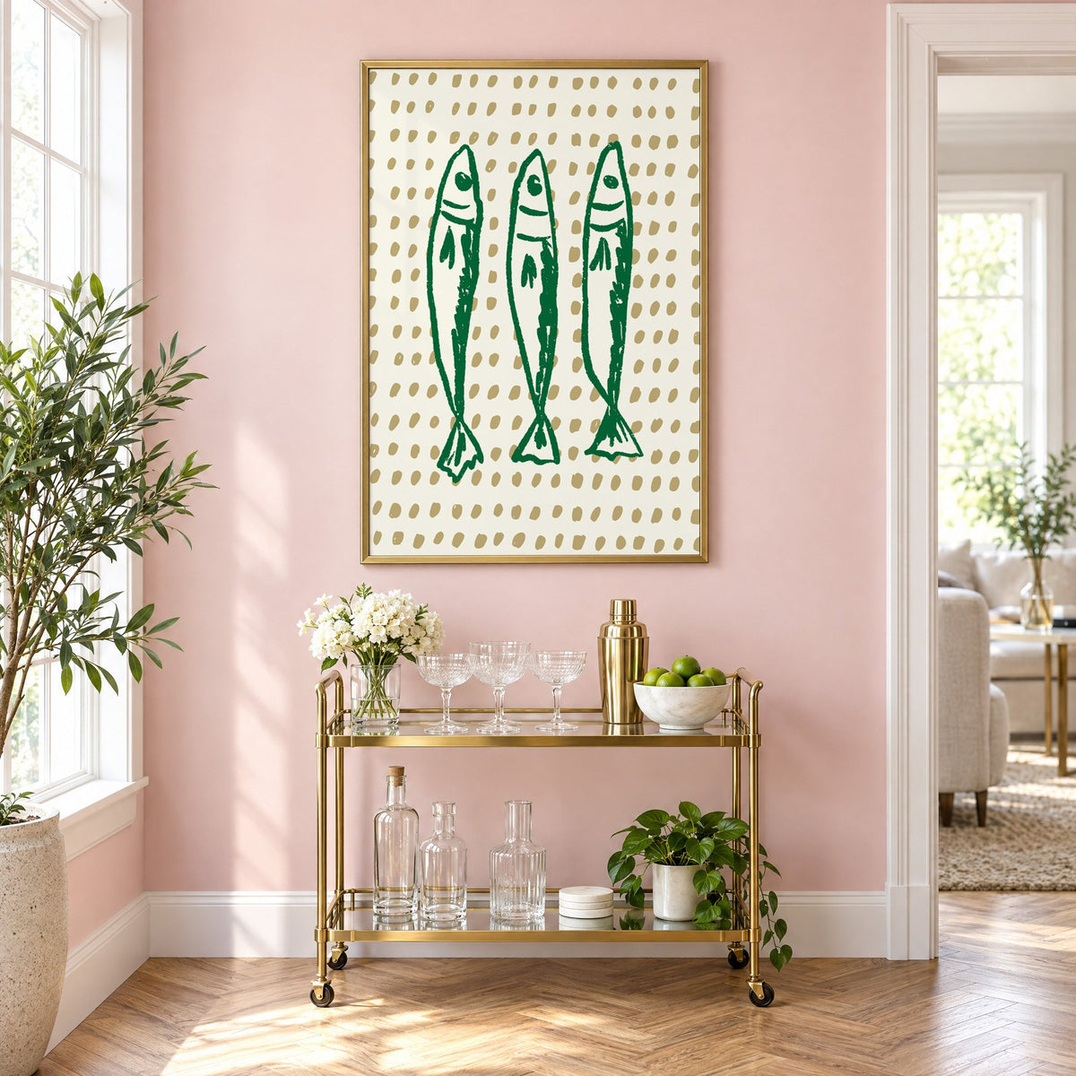 Three green sardines on dotted print in a brass frame on a blush pink wall above a gold bar cart with glassware