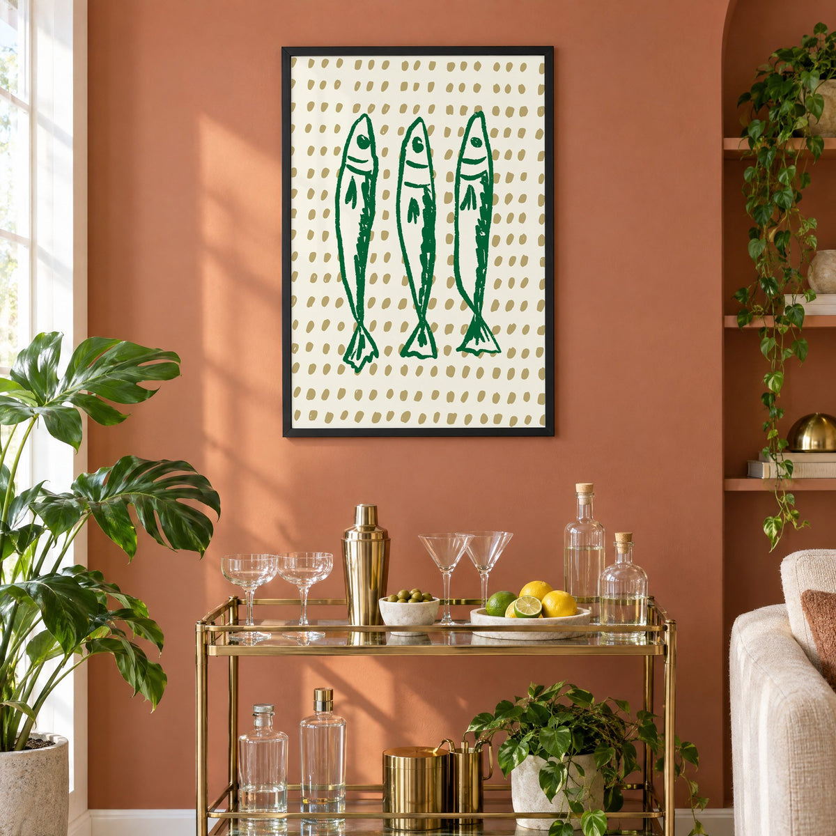 Three green sardines on dotted print in a black frame on a terracotta wall above a brass bar cart with coupes and citrus