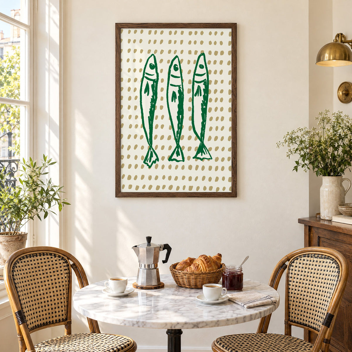 Three green sardines on dotted print in a walnut frame above a marble bistro table with croissants and rattan chairs